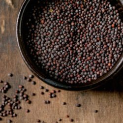 Black mustard seeds for cooking dijon mustard in black ceramic bowl on dark wooden table. Healthy eating, spicy cooking ingredients, traditional kitchen. Top view, close up